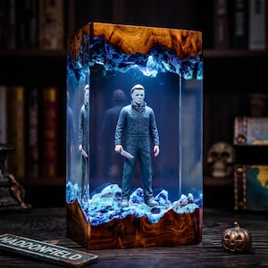 Peut inclure: Une lampe rectangulaire en résine et bois contenant une figurine de Michael Myers. Le personnage porte une combinaison bleu foncé et tient un couteau blanc. La base et le sommet sont en bois naturel, le centre est en résine avec un éclairage bleu.