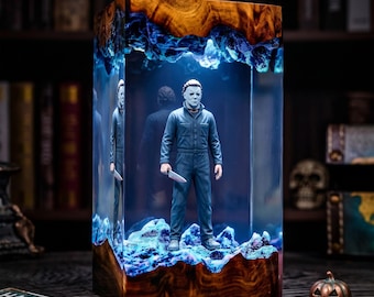 Peut inclure: Une lampe rectangulaire en résine et bois contenant une figurine de Michael Myers. Le personnage porte une combinaison bleu foncé et tient un couteau blanc. La base et le sommet sont en bois naturel, le centre est en résine avec un éclairage bleu.