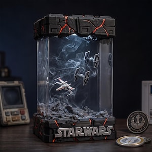 Puede incluir: Una vitrina rectangular con un diorama de Star Wars que incluye un X-wing en miniatura y dos cazas TIE suspendidos en humo. La base y la parte superior son negras con grietas rojas brillantes, y la base muestra el texto plateado STAR WARS.