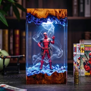 May include: A rectangular resin and wood lamp featuring a Deadpool action figure standing inside. The base and top are made of natural wood, while the centre is clear resin with blue lighting and swirling white smoke effects surrounding the figure.