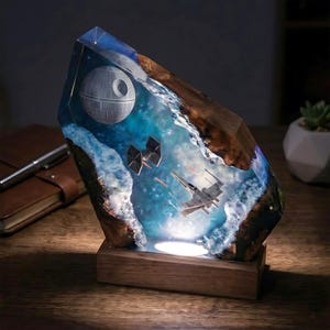May include: A decorative lamp featuring a resin and wood block with a space scene. Inside the translucent blue resin, a spherical space station, a TIE fighter, and an X-wing starfighter are suspended. The base is made of solid wood.