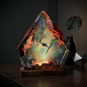 Puede incluir: Una lámpara decorativa de resina con un modelo de caza TIE suspendido dentro de un bloque angular transparente. El interior muestra un efecto de nebulosa en tonos verdes, azules y naranjas, enmarcado por bordes de madera rojiza sobre una base.