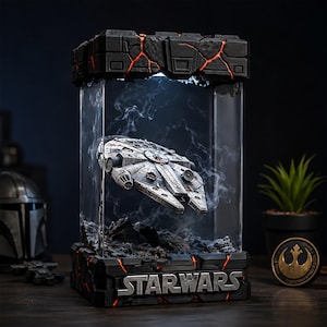 Puede incluir: Un modelo del Halcón Milenario de Star Wars suspendido dentro de una vitrina rectangular transparente. La base y la parte superior tienen paneles negros texturizados con grietas naranjas brillantes. La palabra STAR WARS está en letras plateadas.