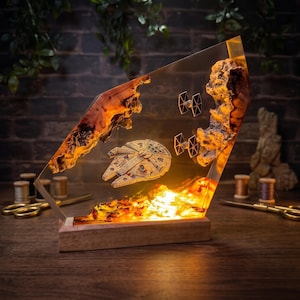 May include: A decorative lamp featuring a Millennium Falcon and two TIE fighters suspended within a clear resin block. The resin is framed by two pieces of natural wood with a glowing orange light at the base, all mounted on a rectangular wooden stand.