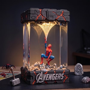 Może przedstawiać: Prostokątna lampa z figurką Spider-Mana w przezroczystej obudowie. Podstawa i góra są ciemnoszare z jarzącymi się pomarańczowymi pęknięciami. Słowo AVENGERS jest srebrne z przodu. Biała pajęczyna rozciąga się od góry do samej figurki.