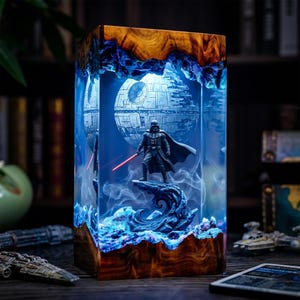 Peut inclure: Une lampe rectangulaire en résine avec une base et un sommet en bois, contenant une figurine miniature de Dark Vador avec un sabre laser rouge, posée sur un rocher devant une sphère représentant l'Étoile de la Mort avec un éclairage bleu.