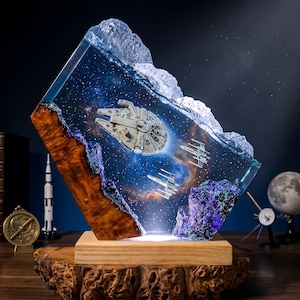 May include: A decorative lamp featuring a resin block with a space scene containing a Millennium Falcon and two X-wing starfighters. The resin is encased by natural wood and purple rock elements, set upon a light-coloured wooden base with internal lighting.