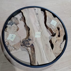 May include: A circular black metal frame containing three distinct pieces of light-colored natural wood with visible grain patterns and knots. Each wood segment features a small, square, light-colored adhesive label with handwritten text.