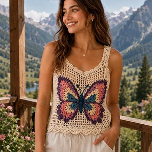 May include: A sleeveless crochet tank top in a cream color featuring a large, multi-colored butterfly motif on the front. The butterfly design incorporates shades of pink, yellow, teal, and dark purple, with a scalloped hem and neckline.
