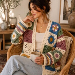 Puede incluir: Un cárdigan de ganchillo de manga larga con un diseño de patchwork de cuadrados de la abuela multicolores. La prenda incorpora motivos florales en tonos azul, verde, amarillo y morado, rematado con un borde acanalado de color crema.
