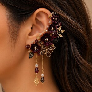 Puede incluir: Un pendiente tipo ear cuff con flores de tela en color burdeos oscuro, hojas de metal dorado y un dije de mariposa dorada. La pieza está decorada con pequeñas perlas blancas y dos cadenas doradas colgantes que terminan en cuentas rojas.