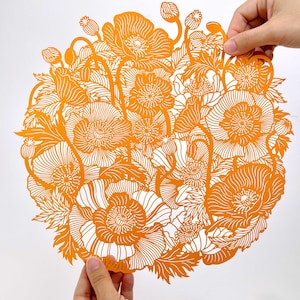 May include: A circular orange paper cutout featuring an intricate, detailed design of blooming poppy flowers, buds, and foliage. The delicate, laser-cut pattern showcases fine lines and organic shapes throughout the entire floral arrangement.