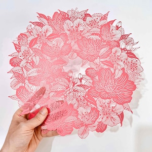 May include: A circular paper cutout featuring an intricate floral pattern in shades of pink and white. The design consists of layered blossoms, buds, and leaves with fine, delicate lines creating a detailed botanical wreath with a central opening.