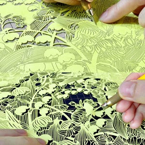 May include: A bright yellow paper sheet featuring an intricate, laser-cut floral pattern of lily of the valley flowers and leaves. A metal craft knife with a textured grip is being used to carefully cut out the delicate, detailed paper design.