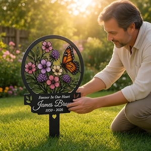 Könnte beinhalten: Ein schwarzer Gedenk-Gartenstecker aus Metall mit einem kreisförmigen Oberteil, das ein buntes Schmetterlings- und Blumenmotiv zeigt. Der Sockel trägt den Text Forever In Our Heart, James Brown, 1950-2026, mit einem kleinen Herzausschnitt.