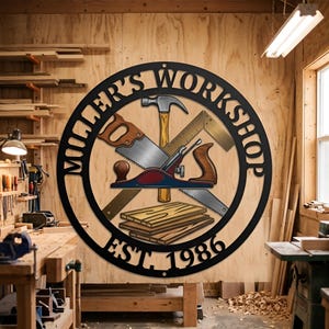 May include: A circular black metal wall sign featuring the text "MILLER'S WORKSHOP" and "EST. 1986". The center displays a graphic illustration of a hammer, a hand saw, a square ruler, a wood plane, and a stack of wooden boards.