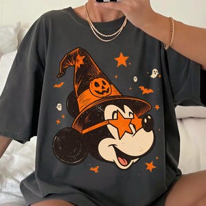 May include: A charcoal gray t-shirt featuring a graphic of a cartoon mouse wearing a witch hat with a pumpkin design and star-shaped orange sunglasses. The print includes small orange stars, bats, and white ghosts scattered around the central character.
