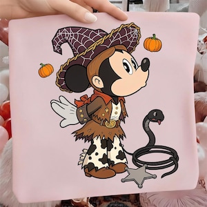May include: A light pink t-shirt featuring a graphic of a cartoon mouse dressed as a cowboy with a witch hat, brown jacket, and cow-print pants. The mouse is accompanied by two orange pumpkins, a black snake, and a silver sheriff star badge.