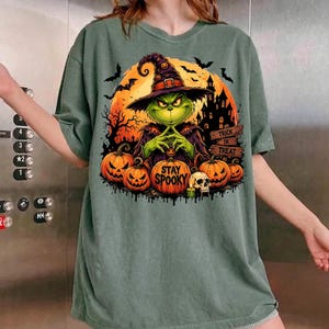 May include: A sage green t-shirt featuring a graphic of a green creature wearing a witch hat, set against a large orange moon. The design includes pumpkins, a skull, a haunted house, bats, and the text "STAY SPOOKY" and "TRICK OR TREAT".