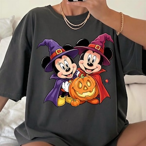May include: A charcoal gray t-shirt featuring a graphic print of two cartoon mice dressed as witches. One mouse wears a purple hat and cape, while the other wears a red hat and cape. They are standing next to a large orange jack-o'-lantern pumpkin.