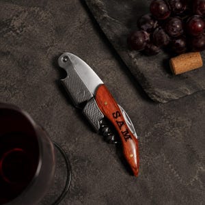 May include: A stainless steel waiter's corkscrew featuring a polished metal body with a textured grip and a wooden handle engraved with the name SAM. The tool includes a folding worm and a foil cutter, presented in a compact, foldable design.