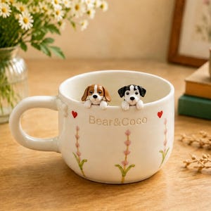 May include: A white ceramic mug featuring two small sculpted dog figures peeking over the rim. The exterior is decorated with painted pink floral sprigs, small red hearts, and the embossed text Bear&Coco in the center of the mug.