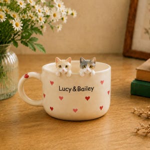 以下が含まれることがあります： 小さな赤いハートと「Lucy & Bailey」の文字がプリントされたクリーム色の陶器製マグカップ。オレンジと白、グレーと白の2匹の猫の彫刻が、マグカップの縁から顔を覗かせています。