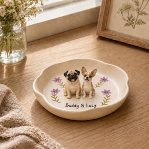 Puede incluir: Un plato decorativo redondo de cerámica color hueso con una figura de un pug y un chihuahua sentados lado a lado en el centro. El plato está decorado con pequeñas flores moradas y el texto impreso Buddy & Lucy bajo los perros.