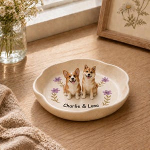 Puede incluir: Un plato decorativo redondo de cerámica color crema con dos pequeñas figuras de perros, un Corgi y un Shiba Inu, en el centro. El plato está decorado con pequeñas ilustraciones de flores moradas y el texto negro "Charlie & Luna".