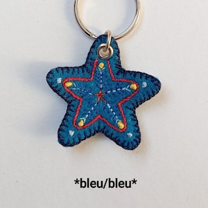 May include: A blue starfish-shaped keyring with a silver ring. The felt keyring has red, white, and yellow embroidered details. The text "*bleu/bleu*" is at the bottom of the image.