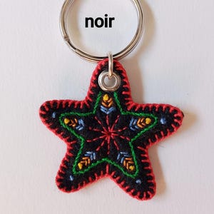 Peut inclure: Un porte-clés en forme d'étoile en tissu noir avec des bords cousus en rouge et un anneau en métal argenté. L'étoile est brodée de motifs verts, jaunes et bleus et porte le mot noir imprimé en noir au-dessus de la forme étoilée.