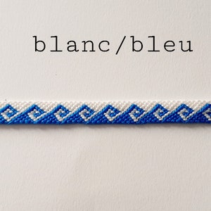 Può includere: Un braccialetto dell'amicizia bianco e blu con un motivo geometrico. Il braccialetto è realizzato con filo da ricamo e ha un semplice design a nodo.