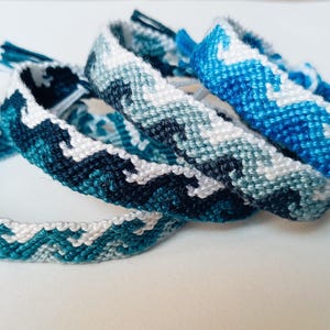 Pode incluir: Quatro pulseiras de amizade feitas à mão em tons de azul e branco. Cada pulseira apresenta um padrão de chevron tecido. As pulseiras são empilhadas, mostrando as diferentes combinações de cores e designs intrincados.