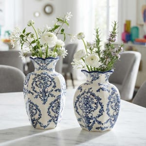 May include: Two ceramic vases with a cream base and intricate blue floral damask patterns. Both vases feature a scalloped rim and a rounded body. The taller vase holds white flowers, while the shorter vase contains white flowers and green herbs.