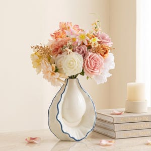 Pode incluir: Um vaso de cerâmica branca com uma silhueta ondulada em forma de rosca com borda azul, contendo um buquê de flores artificiais em tons de pêssego, rosa, creme e amarelo. O vaso está ao lado de uma pilha de três livros e uma vela branca.