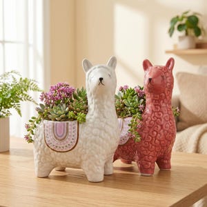 May include: Two ceramic llama-shaped planters, one white and one dusty rose, each featuring a textured wool-like surface and a decorative saddle pattern on the side. Both planters are filled with a variety of small green and pink succulents.