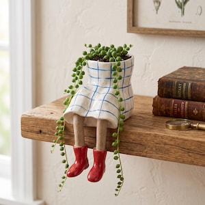 May include: A white ceramic planter shaped like a seated figure wearing a blue grid-patterned skirt and bright red boots. The planter holds a trailing string of pearls plant with small green spherical leaves that drape over the sides.