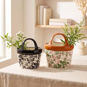 May include: Two ceramic basket-shaped planters with arched handles. The left planter features a black floral pattern on a cream base with a solid black rim. The right planter displays a green and pink floral pattern with a solid brown rim.