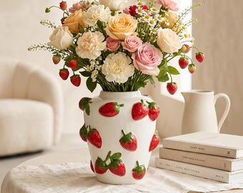 May include: A white ceramic vase featuring raised, three-dimensional red strawberry decorations, filled with a vibrant bouquet of peach, pink, and white roses, carnations, and small white daisies, accented with green stems and small red strawberries.