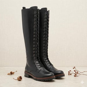 May include: A pair of knee-high black boots featuring a full-length front lace-up closure with silver-toned eyelets. The boots have a rounded toe, decorative brogue-style perforations, a low block heel, and a dark brown stacked sole edge.