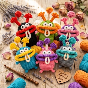 May include: Six colorful crocheted monster toys with large white eyes, wide mouths, and protruding antennae. Each creature features a unique color combination, including black, green, purple, blue, pink, and gray. A wooden heart sign reads Crochet Pattern PDF.