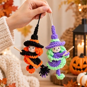 Op de afbeelding: Twee gehaakte heksenornamenten hangend aan touwtjes. De ene heks draagt een zwarte hoed en oranje jurk met oranje haar, terwijl de andere een paarse hoed en groene jurk met groen haar draagt. Beide figuren houden kleine houten bezemstelen vast.
