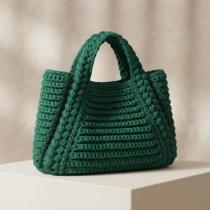 Può includere: Una borsa a mano verde scuro realizzata in filato spesso e testurizzato. La borsa presenta un motivo a coste orizzontali sul corpo principale e due manici integrati con un design intrecciato. La struttura è rigida, con una base ampia.