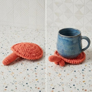 Puede incluir: Un posavasos de ganchillo color coral con forma de tortuga, con un caparazón circular y una pequeña cabeza que sobresale por un lado. El posavasos se muestra solo y con una taza de cerámica azul sobre su superficie tejida a mano.