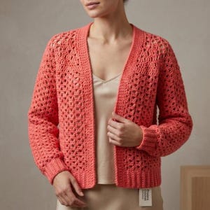 May include: A coral-colored open-front cardigan featuring an intricate crochet pattern with long sleeves and ribbed cuffs. The garment has a V-neckline and a straight hem. A small white rectangular tag with the text Prairie Crochet is attached.