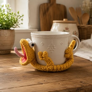 May include: A white ceramic mug featuring a textured, embossed pattern around the rim and body, wrapped in a yellow crocheted lizard cozy. The lizard has a wide-open mouth with a pink tongue, two white eyes with black pupils, and a long tail.