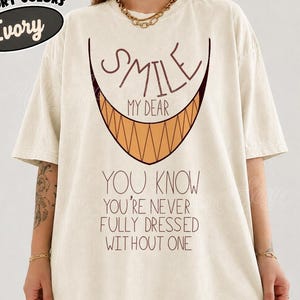 May include: An ivory short-sleeve t-shirt featuring a graphic of a wide, sharp-toothed grin in orange and brown. The text reads: SMILE MY DEAR YOU KNOW YOU'RE NEVER FULLY DRESSED WITHOUT ONE. The shirt has a crew neckline and a relaxed, loose fit.