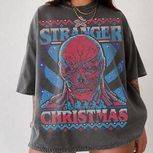 May include: A charcoal gray short-sleeve t-shirt featuring a graphic print with a red creature face and the text STRANGER CHRISTMAS. The design incorporates a blue and white knitted pattern style with geometric shapes and decorative borders.