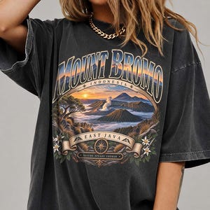 May include: A charcoal gray short-sleeve t-shirt featuring a graphic print of Mount Bromo in East Java, Indonesia. The design includes a sunrise landscape, a compass icon, and the text "MOUNT BROMO," "INDONESIA," "EAST JAVA," and "NATURE SPEAKS LOUDER."