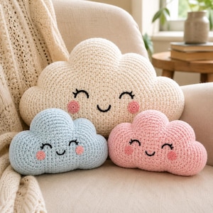 May include: Three cloud-shaped crochet pillows with smiling faces. The largest pillow is cream-colored, while the two smaller pillows are light blue and light pink. Each pillow features embroidered black eyes, a black mouth, and round pink cheeks.